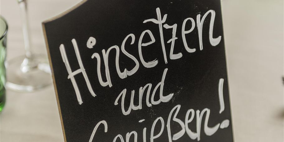 Ein schwarzes Schild mit der Aufschrift "Hinsetzen und Genießen!" steht auf einem Tisch. Um das Schild sind Gläser in verschiedenen Farben angeordnet.