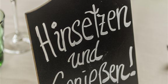 A black sign with the inscription "Sit Down and Enjoy!" stands on a table. Glasses in various colors are arranged around the sign.
