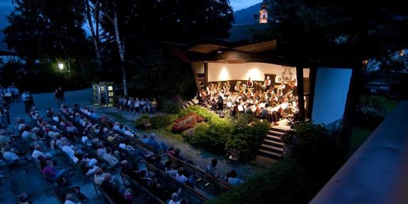 Ein Konzert im Freien mit einem Orchester auf einer Bühne. Viele Zuschauer sitzen auf Bänken und genießen die Musik in einer gemütlichen Abendstimmung.