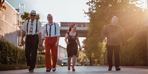 Vier Menschen gehen gemeinsam auf einer Straße, umgeben von Bäumen und Gebäuden. Die Sonne scheint und schafft eine warme Atmosphäre.