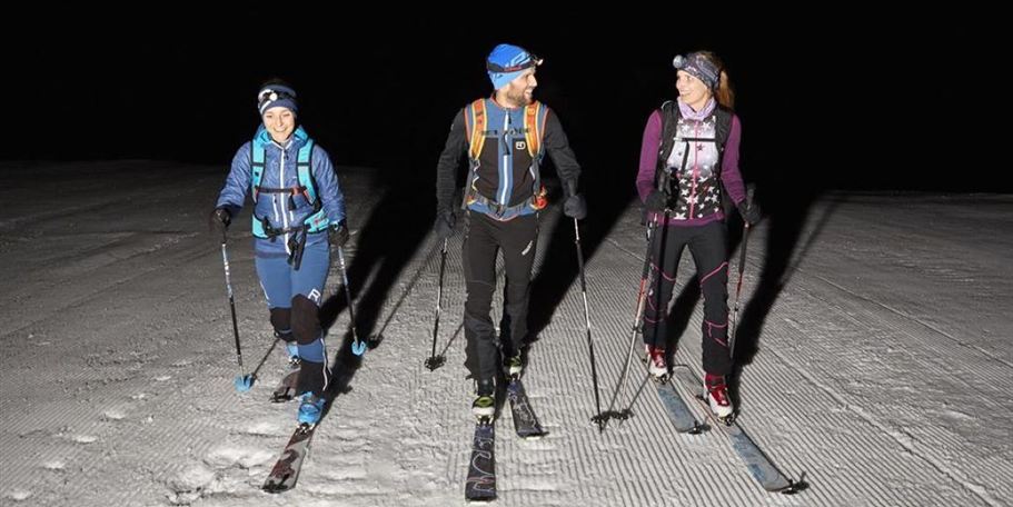 Three skiers are walking at night over a snow-covered slope. They are wearing headlamps and skiing in a deep, dark environment.