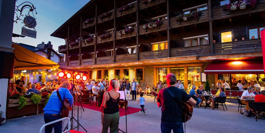 Eine belebte Straßenszene mit Musikern, die vor einem Holzgebäude auftreten. Viele Menschen sitzen draußen und genießen die Atmosphäre am Abend.