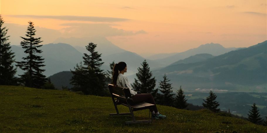 Eine Person sitzt alleine auf einer Bank und schaut auf eine malerische Berglandschaft während des Sonnenuntergangs. Die Szene ist von Bäumen umgeben und vermittelt eine ruhige, entspannende Atmosphäre.