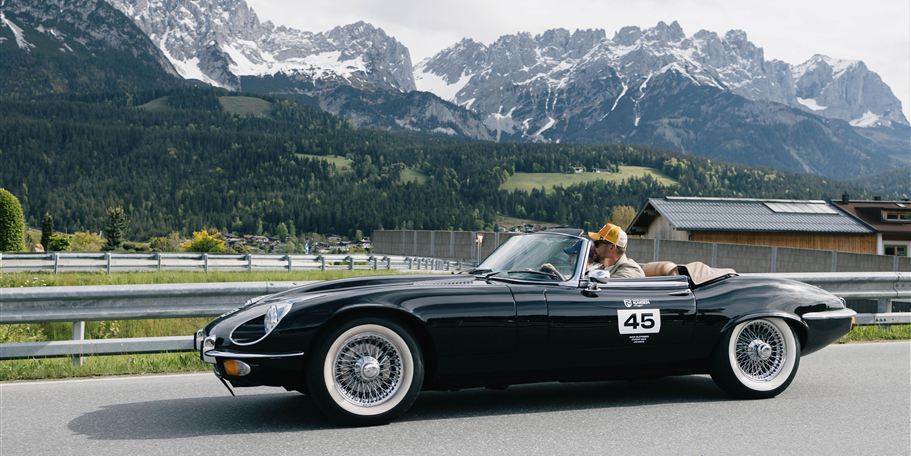 A black convertible drives along a winding road with impressive mountains in the background. The driver is wearing a hat and enjoying the ride in nature.