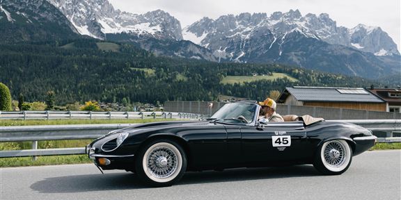 A black convertible drives along a winding road with impressive mountains in the background. The driver is wearing a hat and enjoying the ride in nature.