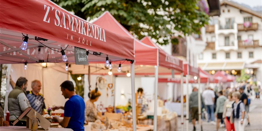 Ein lebendiger Marktplatz bei Dämmerung mit Ständen, die lokale Produkte anbieten. Menschen schlendern durch die Gassen und genießen die Atmosphäre.