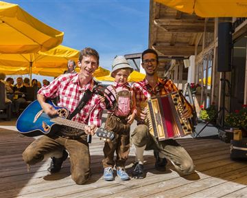 Two musicians in traditional clothing are playing with a child. They are standing on a terrace with yellow umbrellas in the background.