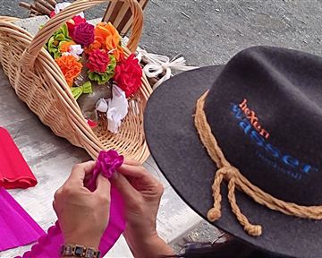 A person is crafting flowers out of colorful fabric. In the background, there is a basket with already completed flowers.