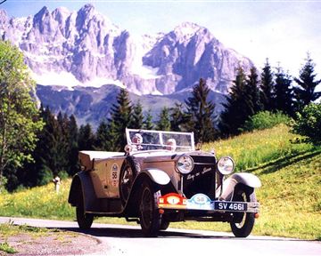 A classic car drives along a picturesque road in the mountains. The surroundings are green with tall trees and impressive mountain peaks in the background.