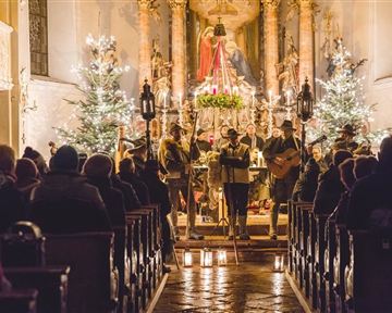 Eine festlich geschmückte Kirche mit Weihnachtsbäumen und Kerzenlicht. Eine Musikgruppe spielt vor den Zuhörern in der schönen Umgebung.