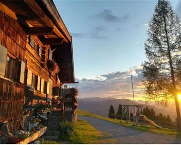 Ein rustikales Holzhaus mit blühenden Pflanzen steht gegen den Sonnenuntergang. Im Hintergrund sind Berge und eine sanfte Hügellandschaft sichtbar.