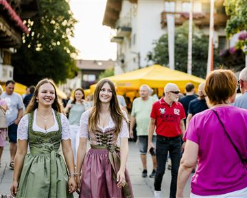 Zwei Frauen in traditionellen Dirndlkleidern gehen fröhlich durch eine belebte Straße. Im Hintergrund sind Stände und viele Menschen zu sehen.