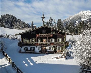 A traditional chalet in the snow, surrounded by fir trees. The sky is blue and cloudy, creating an idyllic winter landscape.