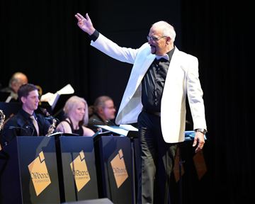 Ein Dirigent in einem weißen Smoking leitet ein Orchester. Im Hintergrund sind Musiker mit ihren Instrumenten zu sehen.