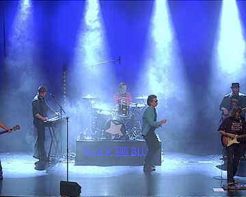A band is playing live on stage with colored lights and fog. The musicians are in vibrant poses and are using various instruments.