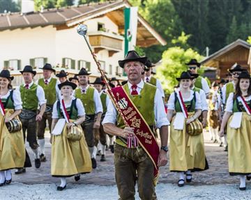 Eine Gruppe von Menschen in traditioneller Tracht marschiert festlich in einer ländlichen Umgebung. Der Anführer trägt einen Stab und eine besondere Auszeichnung.