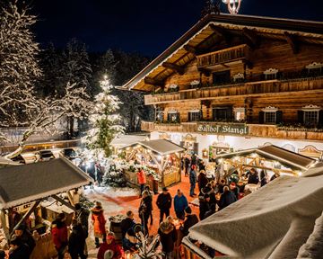 A festive winter market scene with people visiting stalls. In the background stands a rustic wooden house, surrounded by snow-covered trees and a lit Christmas tree.