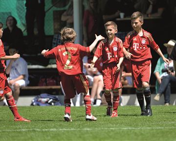 Eine Gruppe von jungen Fußballspielern feiert ein Tor auf dem Spielfeld. Sie tragen rot-weiße Trikots und wirken glücklich und aufgeregt.