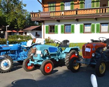 Three historic tractors stand in front of a traditional country house. The tractors are painted in various colors and display nostalgic charm.