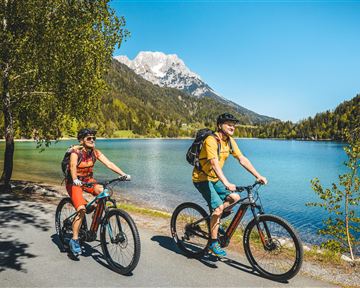Zwei Radfahrer fahren an einem klaren blauen See entlang, umgeben von Bergen und Bäumen. Die Sonne scheint und der Himmel ist blau.