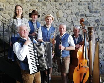 Six musicians in traditional attire stand in front of a stone wall. They hold various instruments such as accordion, clarinet, and harp.