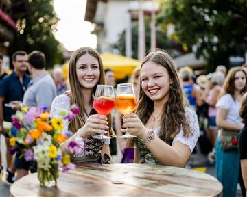 Zwei junge Frauen stoßen mit bunten Getränken an. Im Hintergrund sind Menschen und Marktstände zu sehen, die eine festliche Atmosphäre erzeugen.