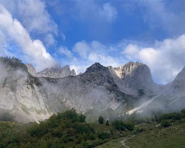 Ein beeindruckendes Bergpanorama mit felsigen Gipfeln und leichten Nebelschwaden. Der Himmel ist klar mit einigen Wolken.