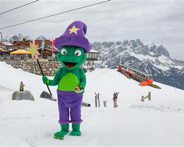 A green wizard in a colorful costume stands in the snow in front of a mountain landscape. In the background, a ski lift and a mountain cabin can be seen.