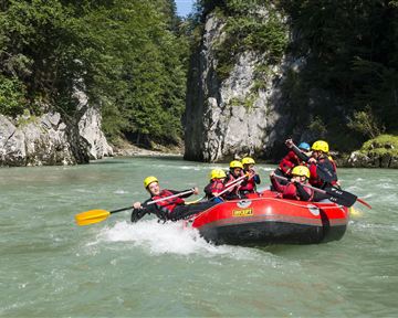 A group of people paddles in a red raft on a clear river. Surrounded by green trees and rocks, the scenery provides an adventurous experience in nature.