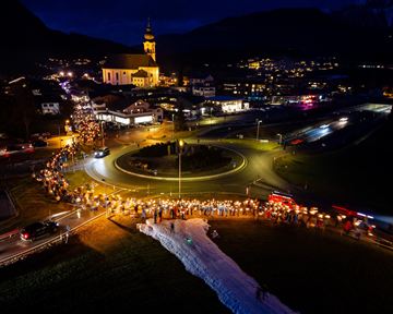 Eine festliche Veranstaltung bei Nacht mit vielen Menschen, die Lichter und Laternen tragen. Im Hintergrund ist eine Kirche und eine kleinräumige Stadtansicht zu sehen.