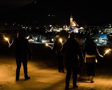 A group of people stands in the snow at night, holding torches in their hands. In the background, a lit city with a church can be seen.