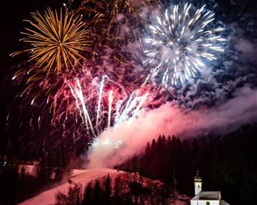 A spectacular fireworks display lights up the night sky above a snowy landscape. In the foreground stands a small church surrounded by trees.