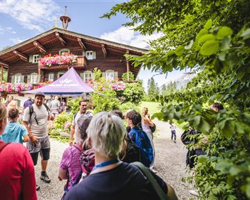 Eine Gruppe von Menschen steht vor einem traditionellen Holzhaus mit blühenden Fenstern. Im Hintergrund ist eine grüne Landschaft zu sehen, die den schönen Tag unterstreicht.