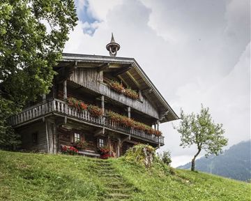 Ein traditionelles Holzhaus mit Balkon und bunten Blumen. Es steht in einer grünen Landschaft unter einem bewölkten Himmel.