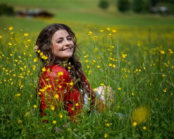 A smiling woman is sitting in a blooming field of yellow flowers. She is wearing a red dress and enjoying nature.