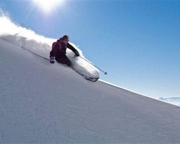 A skier glides down the snowy slope with momentum. The sky is clear and sunny, and fresh snow is being kicked up.