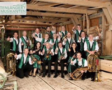 A group of musicians in traditional costume stands in a rustic setting. They hold instruments and smile at the camera.