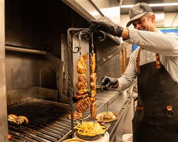 Ein Koch steht am Grill und bereitet Fleisch auf einem Spieß zu. Im Vordergrund sind Pommes frites und ein Burger zu sehen.