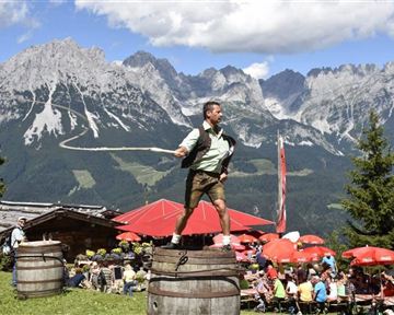 Ein Mann in traditioneller Tracht steht auf einem Fass und schwingt eine Peitsche. Im Hintergrund sind beeindruckende Berge und eine festliche Atmosphäre mit vielen Gästen zu sehen.