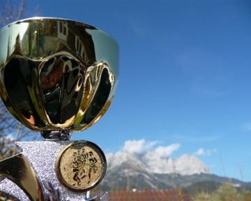 A golden trophy stands in the foreground, with an impressive mountain landscape in the background. The sky is clear and blue.