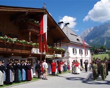 Eine traditionelle Feier in einem malerischen Dorf. Menschen in festlicher Tracht stehen zusammen, umgeben von historischen Gebäuden und Bergen.