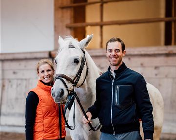 Eine Frau und ein Mann stehen mit einem weißen Pferd in einem Reitstall. Der Mann trägt eine dunkle Jacke und die Frau eine orange Weste.