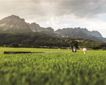 A golf course with green grass and a golf ball on a tee. In the background, impressive mountains and a cloudy sky can be seen.