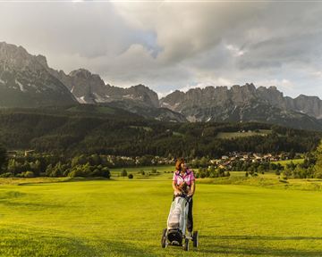 A woman with a golf cart is standing on a green golf course. Impressive mountains and a cloudy sky can be seen in the background.