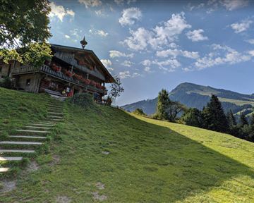 Ein malerisches Landhaus auf einer grünen Wiese mit sanften Hügeln im Hintergrund. Der Himmel ist klar und blau, mit einigen Wolken.