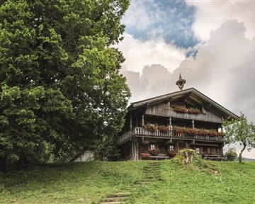Ein traditionelles Holzhaus steht auf einem sanften Hügel, umgeben von Bäumen. Der Himmel ist bewölkt, was eine ruhige Atmosphäre schafft.
