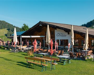 Ein gemütliches Restaurant in der Alpenlandschaft mit einer großen Terrasse. Umgeben von grünen Wiesen und bewaldeten Hügeln.