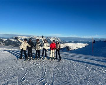 Five skiers are standing on a snowy slope. In the background, there are mountains and a clear blue sky.
