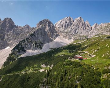 Eine beeindruckende Berglandschaft mit hohen Felsen und grünen Wiesen. Im Vordergrund befindet sich ein rotes Haus in der idyllischen Natur.