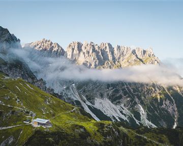 Eine beeindruckende Berglandschaft mit hohen Gipfeln und wolkenbedeckten Hängen. Im Vordergrund ist eine gemütliche Hütte zu sehen.